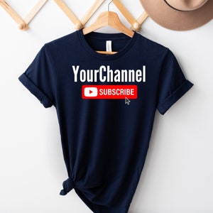 May include: A navy blue t-shirt with the text "YourChannel" and a red YouTube subscribe button with a white play button and the word "SUBSCRIBE" in white text.