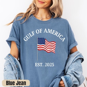 Könnte beinhalten: Hellblaues T-Shirt mit dem Text "GULF OF AMERICA" und einer amerikanischen Flagge. Unter der Flagge steht "EST. 2025". Das T-Shirt hat eine hellblaue Farbe.