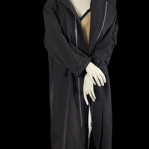 May include: A long, black hooded coat with a zipper closure and a button detail. The coat is displayed on a mannequin, showcasing its sleek design and tailored fit. The coat has a long length, reaching the mid-calf.