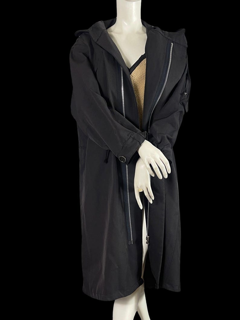 May include: A long, black hooded coat with a zipper closure and a button detail. The coat is displayed on a mannequin, showcasing its sleek design and tailored fit. The coat has a long length, reaching the mid-calf.