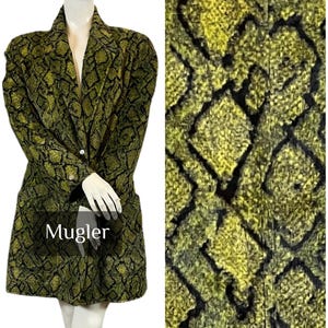 Peut inclure: Un manteau à motifs verts et noirs avec le texte "Mugler". Le manteau a une coupe longue et ajustée avec un col cranté et une fermeture boutonnée. Le motif ressemble à une peau de serpent.
