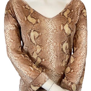 Puede incluir: Una blusa de manga larga con cuello en V, con un estampado de piel de serpiente en tonos marrón y beige. La blusa se muestra en un maniquí, mostrando su diseño y ajuste. Las mangas están dobladas en las muñecas.