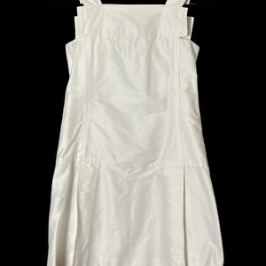May include: A white dress with a square neckline and wide shoulder straps. The dress has a fitted bodice and a flared skirt with a front pleat. The dress is made of a smooth, shiny fabric and hangs on a black hanger.