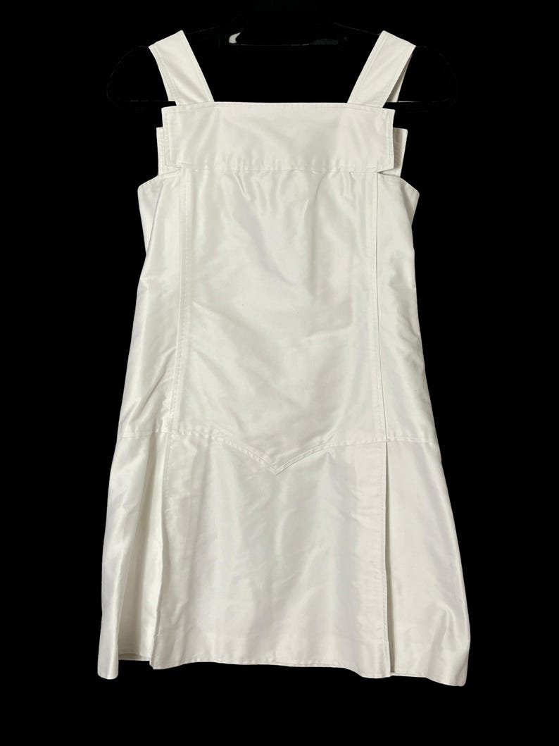 May include: A white dress with a square neckline and wide shoulder straps. The dress has a fitted bodice and a flared skirt with a front pleat. The dress is made of a smooth, shiny fabric and hangs on a black hanger.