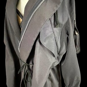 May include: Black trench coat with a draped collar, a full-length zipper, and a drawstring waist. The coat features long sleeves with elastic cuffs and a small zippered pocket on the upper arm. The fabric has a slight sheen.