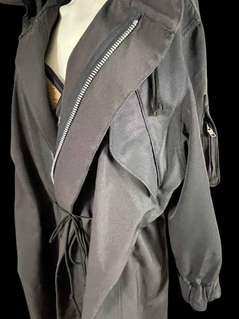 May include: Black trench coat with a draped collar, a full-length zipper, and a drawstring waist. The coat features long sleeves with elastic cuffs and a small zippered pocket on the upper arm. The fabric has a slight sheen.