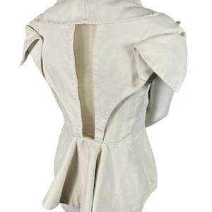 以下が含まれることがあります： ユニークなデザインのクリーム色の構造化されたジャケット。ハイカラー、建築的なショルダーディテール、ペプラムスタイルのウエストが特徴です。背面には垂直の切り抜きがあります。マネキンに展示されています。
