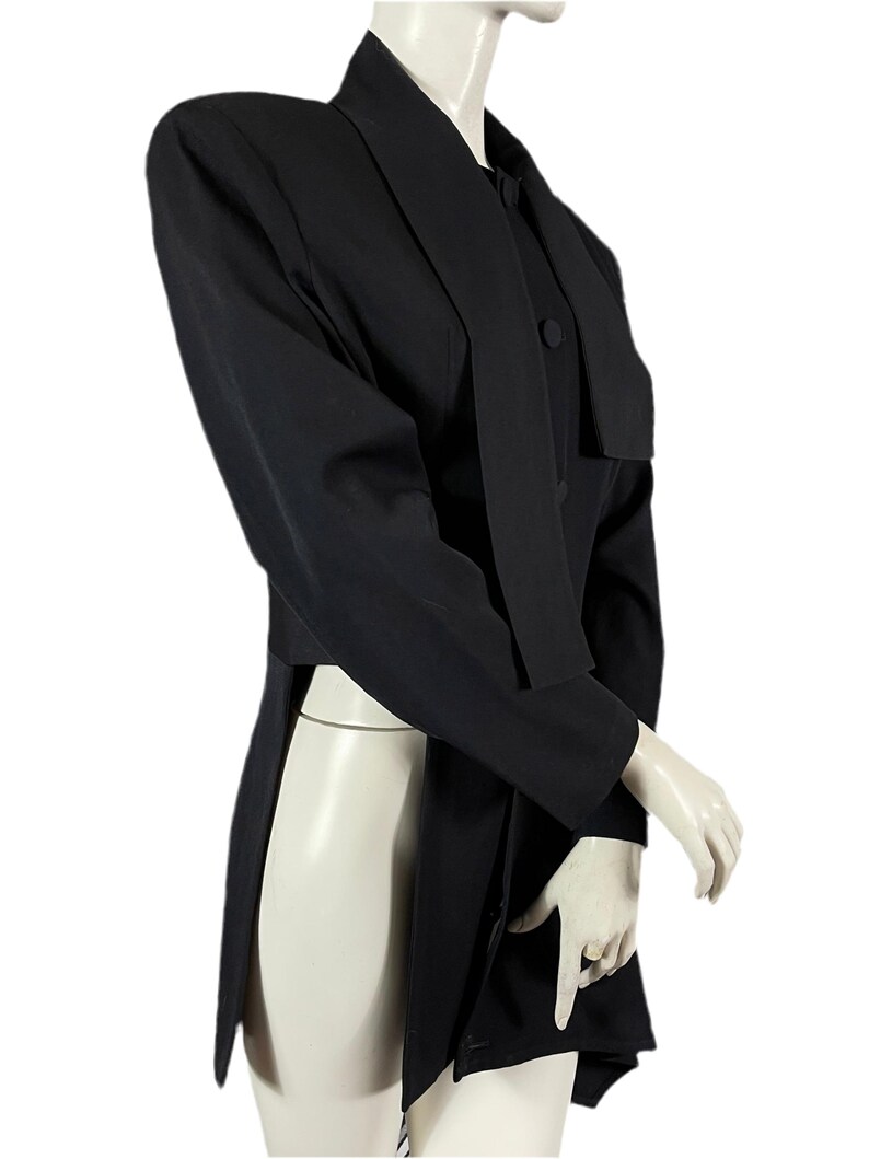 May include: A black blazer with a unique design, featuring a long, flowing panel on the front and a notched lapel. The blazer has long sleeves and a button closure. The garment is displayed on a mannequin.
