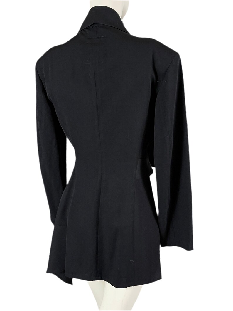 May include: A black blazer with long sleeves, a notched collar, and a flared hem. The back view of the blazer shows a tailored fit, emphasizing the waist. The garment is displayed on a mannequin against a white background.