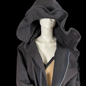 May include: A dark gray hooded jacket with a large, flowing hood. The jacket features a full-length zipper and a contrasting tan lining. The hood is designed to drape over the shoulders, creating a unique silhouette. The jacket is displayed on a mannequin.