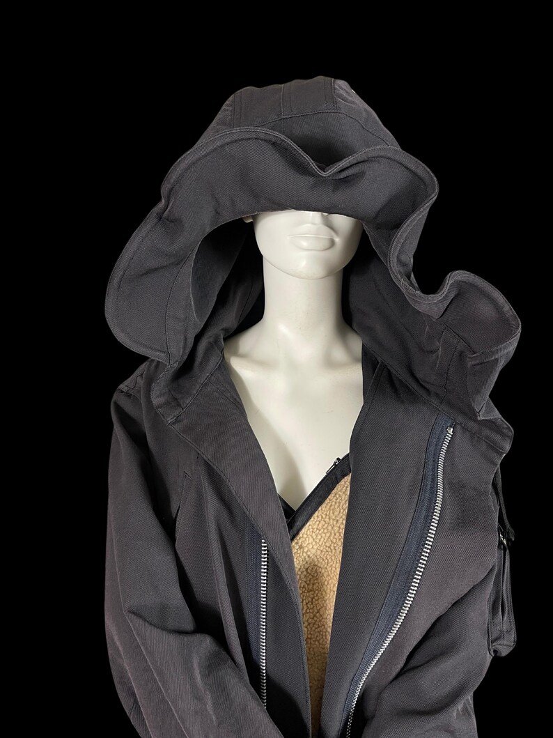 May include: A dark gray hooded jacket with a large, flowing hood. The jacket features a full-length zipper and a contrasting tan lining. The hood is designed to drape over the shoulders, creating a unique silhouette. The jacket is displayed on a mannequin.