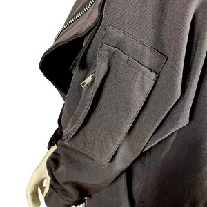 May include: Close-up of a black jacket sleeve with a zippered pocket and a smaller zippered compartment. The jacket has a textured fabric and a visible zipper detail along the upper arm. The sleeve is on a mannequin.