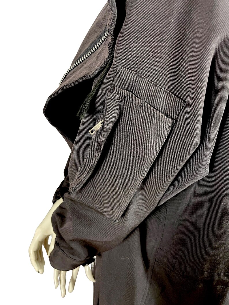 May include: Close-up of a black jacket sleeve with a zippered pocket and a smaller zippered compartment. The jacket has a textured fabric and a visible zipper detail along the upper arm. The sleeve is on a mannequin.