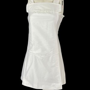 May include: A white sleeveless dress with a square neckline and wide shoulder straps. The dress features a unique design element on the front, with a V-shaped detail at the hem. The dress is displayed on a mannequin against a black background.