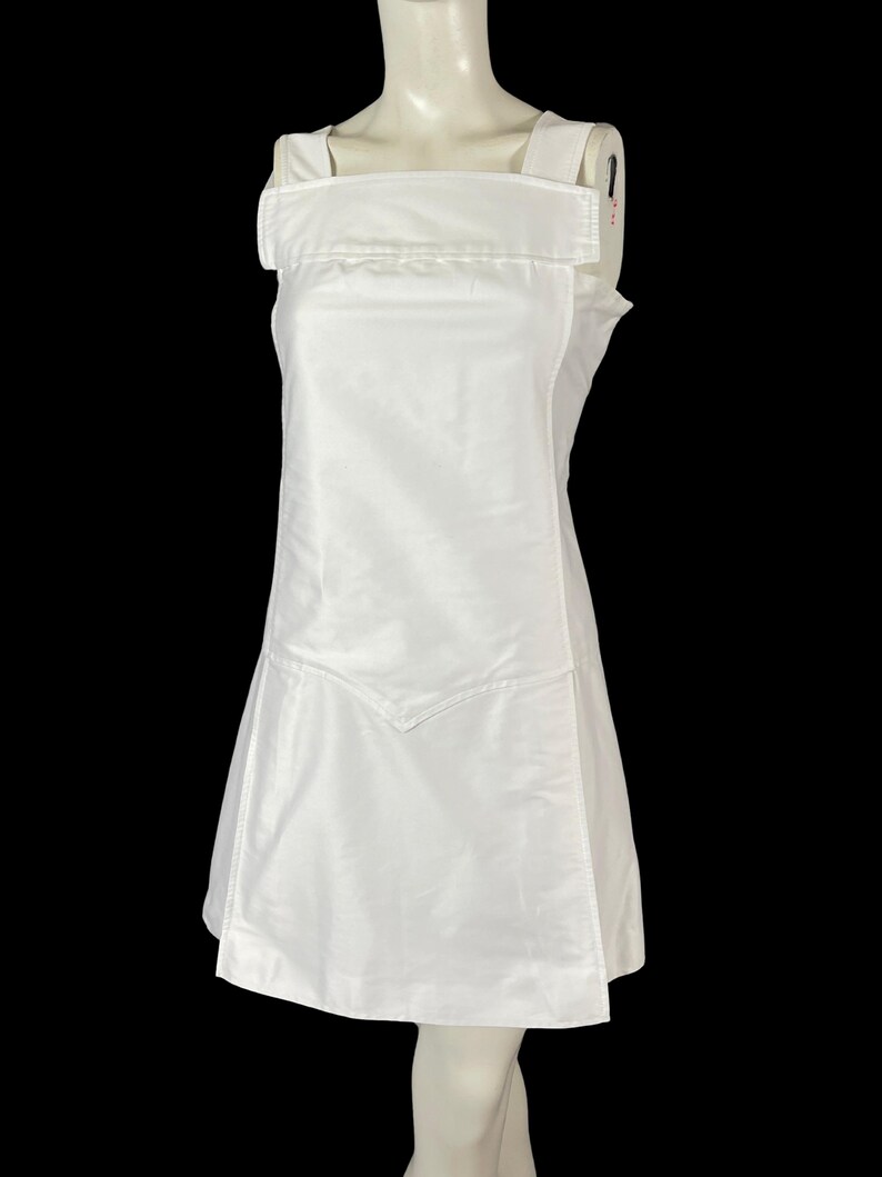May include: A white sleeveless dress with a square neckline and wide shoulder straps. The dress features a unique design element on the front, with a V-shaped detail at the hem. The dress is displayed on a mannequin against a black background.