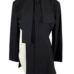 May include: A black long-sleeved jacket with a unique design. The jacket features a high collar that extends down the front, a button-up closure, and an asymmetrical hemline. The jacket is displayed on a mannequin.