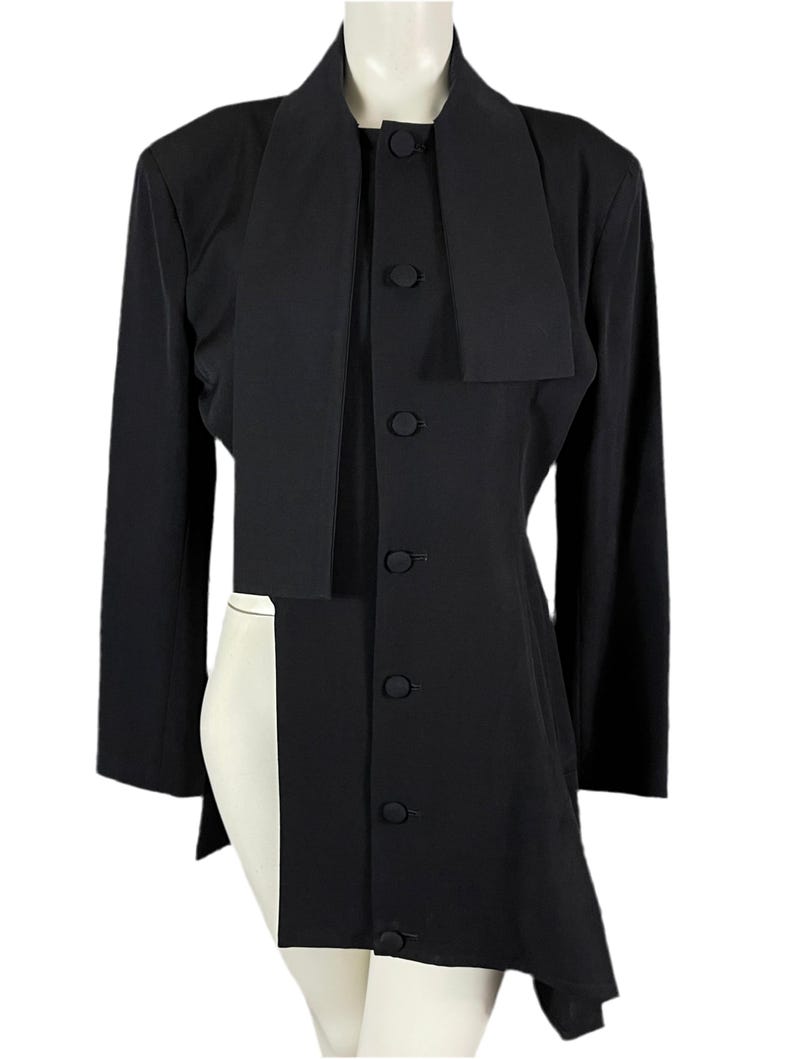 May include: A black long-sleeved jacket with a unique design. The jacket features a high collar that extends down the front, a button-up closure, and an asymmetrical hemline. The jacket is displayed on a mannequin.