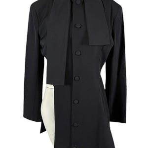 May include: A black long-sleeved jacket with a unique design. The jacket features a high collar, a button-down front, and an asymmetrical hemline. The jacket has a long, draped panel that extends down the front. The jacket is made of a smooth, solid black fabric.
