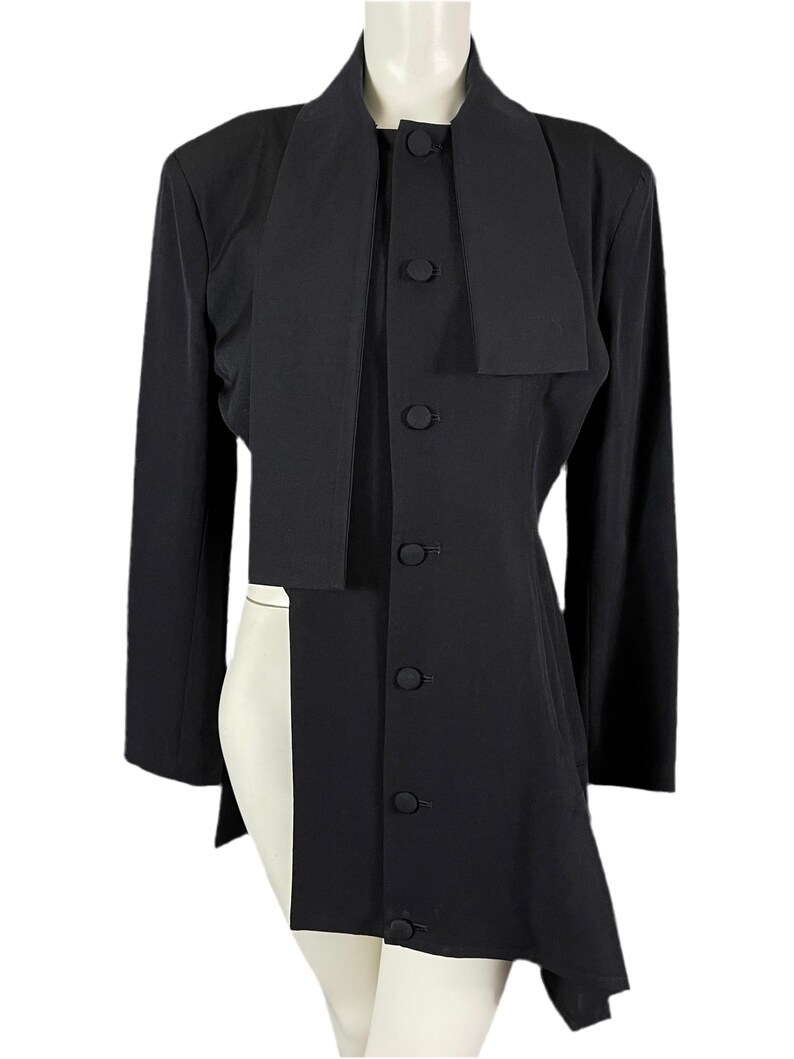 May include: A black long-sleeved jacket with a unique design. The jacket features a high collar, a button-down front, and an asymmetrical hemline. The jacket has a long, draped panel that extends down the front. The jacket is made of a smooth, solid black fabric.