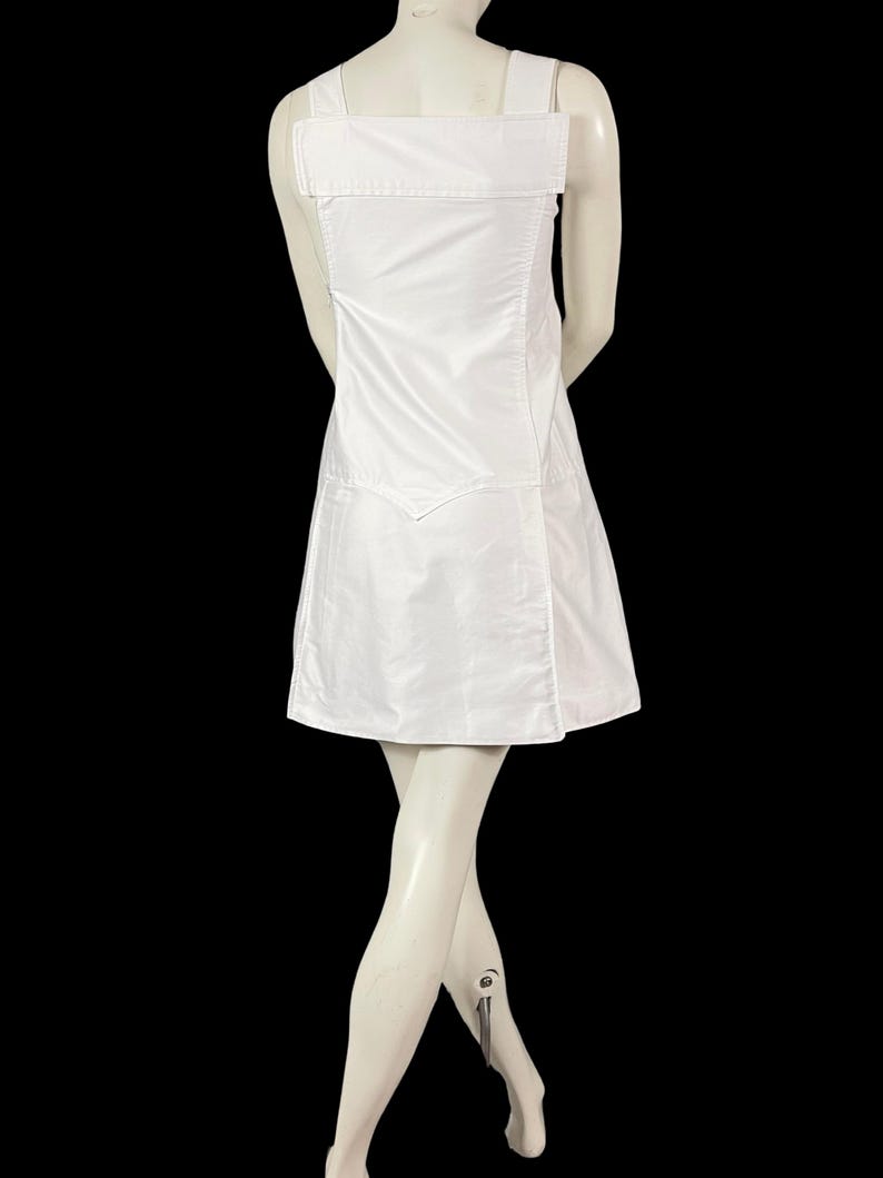 May include: A white dress with a square neckline and wide shoulder straps. The dress has a fitted bodice and a flared skirt. The back of the dress features a large, rectangular panel. The dress is displayed on a mannequin against a black background.