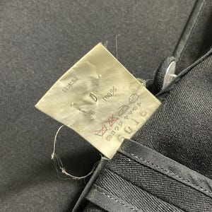 May include: Close-up of black fabric with a tag attached. The tag has Japanese text and symbols, including "100%" and "日本製". The fabric appears to be a dark, possibly denim material, with visible stitching.