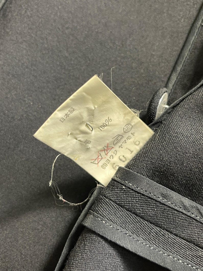 May include: Close-up of black fabric with a tag attached. The tag has Japanese text and symbols, including "100%" and "日本製". The fabric appears to be a dark, possibly denim material, with visible stitching.