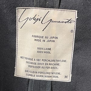 May include: Close-up of a garment label with the designer's name, "Yohji Yamamoto," in elegant script. The label indicates the item is made in Japan from 100% wool. Care instructions include dry cleaning and warm iron. The size is M.