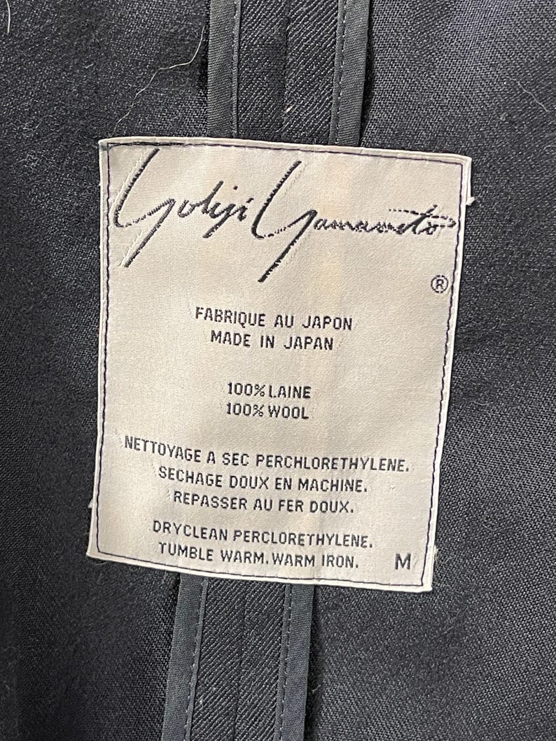 May include: Close-up of a garment label with the designer's name, "Yohji Yamamoto," in elegant script. The label indicates the item is made in Japan from 100% wool. Care instructions include dry cleaning and warm iron. The size is M.