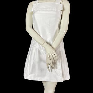 May include: A white sleeveless dress with a square neckline and a-line skirt. The dress features wide shoulder straps and a fitted bodice. The dress is displayed on a mannequin against a black background. The dress appears to be made of a smooth, possibly satin-like fabric.