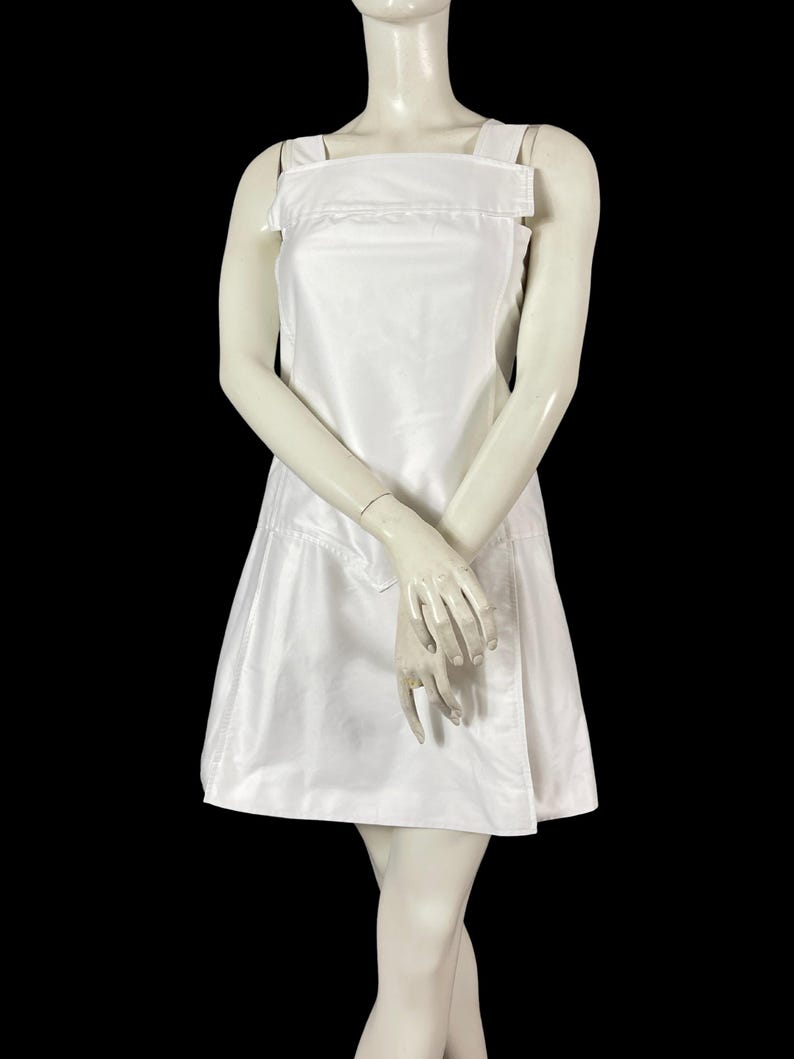 May include: A white sleeveless dress with a square neckline and a-line skirt. The dress features wide shoulder straps and a fitted bodice. The dress is displayed on a mannequin against a black background. The dress appears to be made of a smooth, possibly satin-like fabric.