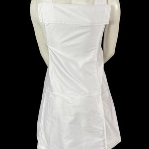 May include: A white dress with a unique design. The dress features wide shoulder straps and a large rectangular panel across the upper back. The skirt portion has a wrap-style design. The dress is made of a smooth, possibly satin-like fabric.