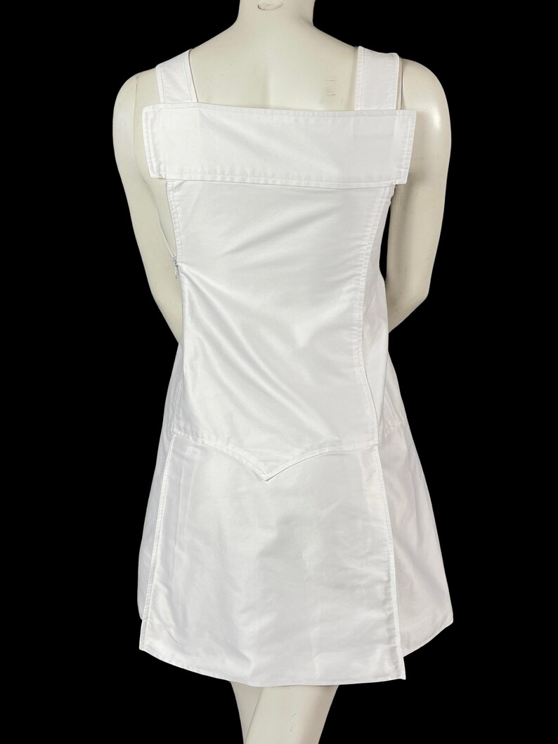 May include: A white dress with a unique design. The dress features wide shoulder straps and a large rectangular panel across the upper back. The skirt portion has a wrap-style design. The dress is made of a smooth, possibly satin-like fabric.