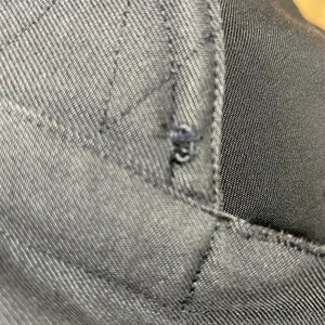 May include: Close-up of dark gray fabric with visible stitching and a small tear. The fabric appears to be a sturdy material, possibly denim or a similar woven textile. The stitching forms a quilted pattern.