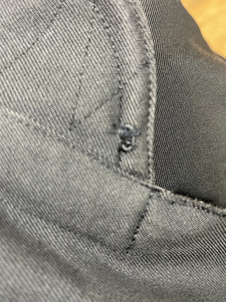 May include: Close-up of dark gray fabric with visible stitching and a small tear. The fabric appears to be a sturdy material, possibly denim or a similar woven textile. The stitching forms a quilted pattern.
