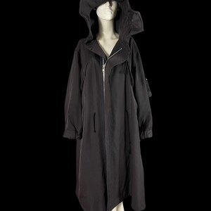 May include: A long, black hooded coat with a zipper closure, displayed on a mannequin. The coat features a large hood and long sleeves. The fabric appears to be a lightweight material. The background is solid black.