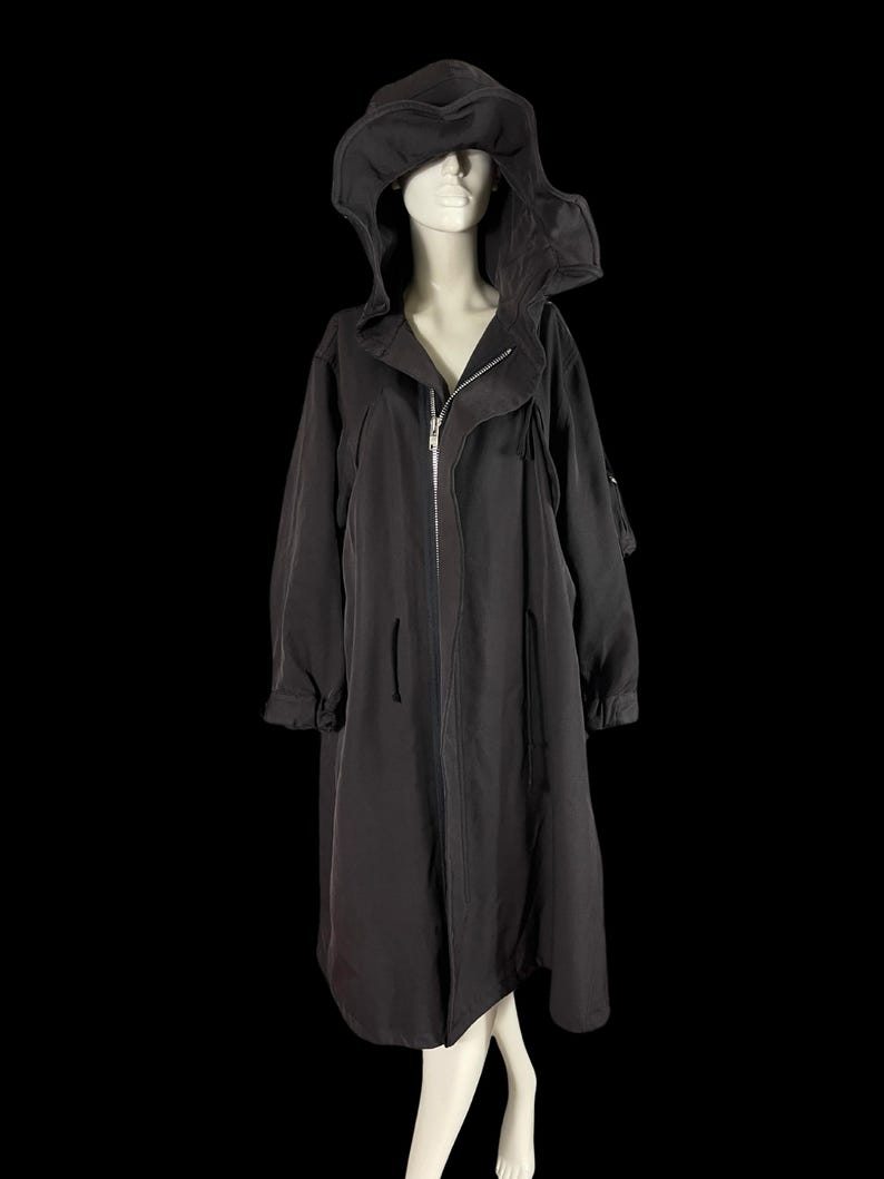 May include: A long, black hooded coat with a zipper closure, displayed on a mannequin. The coat features a large hood and long sleeves. The fabric appears to be a lightweight material. The background is solid black.