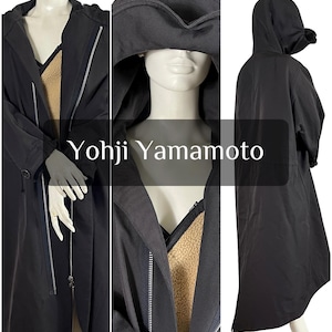 May include: Black long coat with a hood and a zipper closure. The coat has a contrasting tan lining and the text "Yohji Yamamoto" is displayed on the front. The coat is displayed on a mannequin.