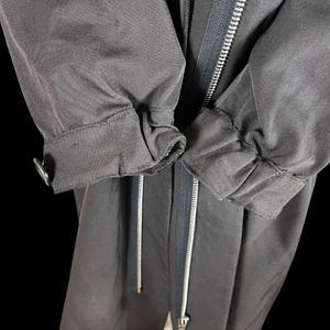 May include: Close-up of a dark gray jacket with a silver zipper and drawstring. The sleeves are gathered at the cuffs with a button closure. The jacket appears to be made of a durable, possibly water-resistant fabric.