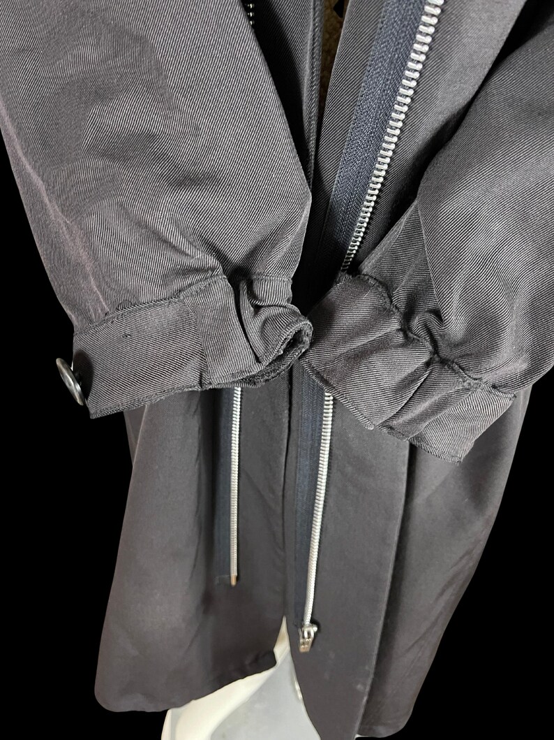 May include: Close-up of a dark gray jacket with a silver zipper and drawstring. The sleeves are gathered at the cuffs with a button closure. The jacket appears to be made of a durable, possibly water-resistant fabric.