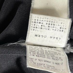 May include: Close-up of a dark gray fabric with attached white tags. The tags feature Japanese and English text, including care instructions and material information. The fabric has a subtle texture.