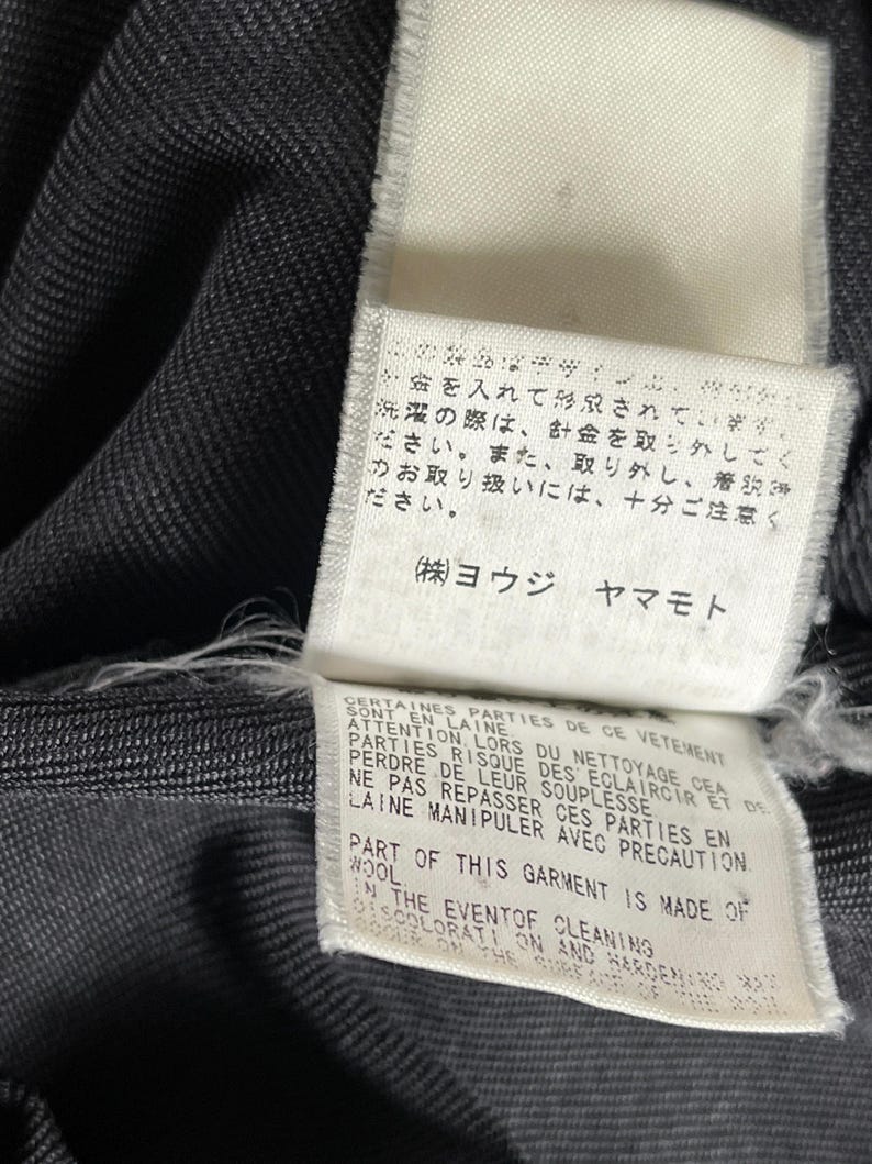 May include: Close-up of a dark gray fabric with attached white tags. The tags feature Japanese and English text, including care instructions and material information. The fabric has a subtle texture.