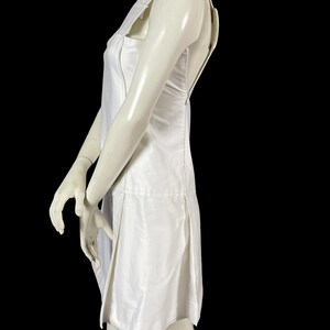 May include: A white sleeveless dress with a square neckline and a knee-length hem. The dress features a fitted bodice and a pleated skirt. The dress is displayed on a mannequin against a black background.