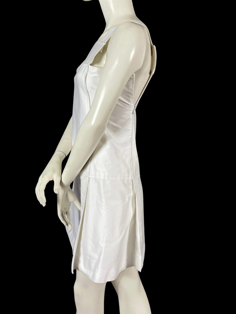May include: A white sleeveless dress with a square neckline and a knee-length hem. The dress features a fitted bodice and a pleated skirt. The dress is displayed on a mannequin against a black background.