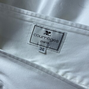 May include: Close-up of a white garment with a label that reads "courr&egrave;ges paris" in black text. The label also has a logo and the number "36". The fabric is smooth and appears to be a light material.