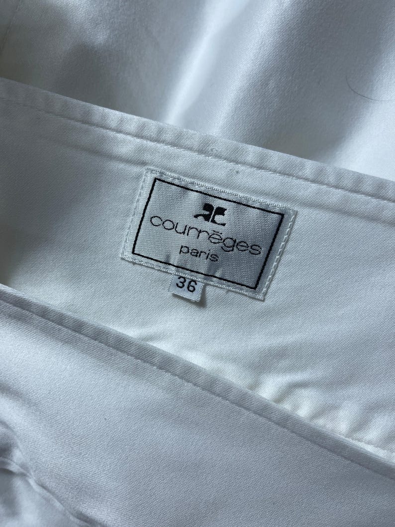 May include: Close-up of a white garment with a label that reads "courr&egrave;ges paris" in black text. The label also has a logo and the number "36". The fabric is smooth and appears to be a light material.