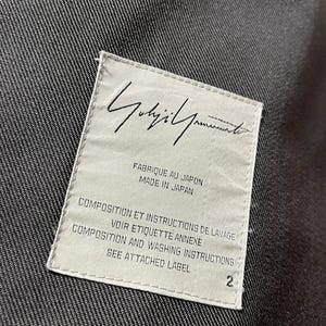 May include: A white label with black text, featuring the brand name "Yohji Yamamoto" in cursive script. The label includes text in English and French, indicating the item's origin and care instructions. The number "2" is printed on the label.