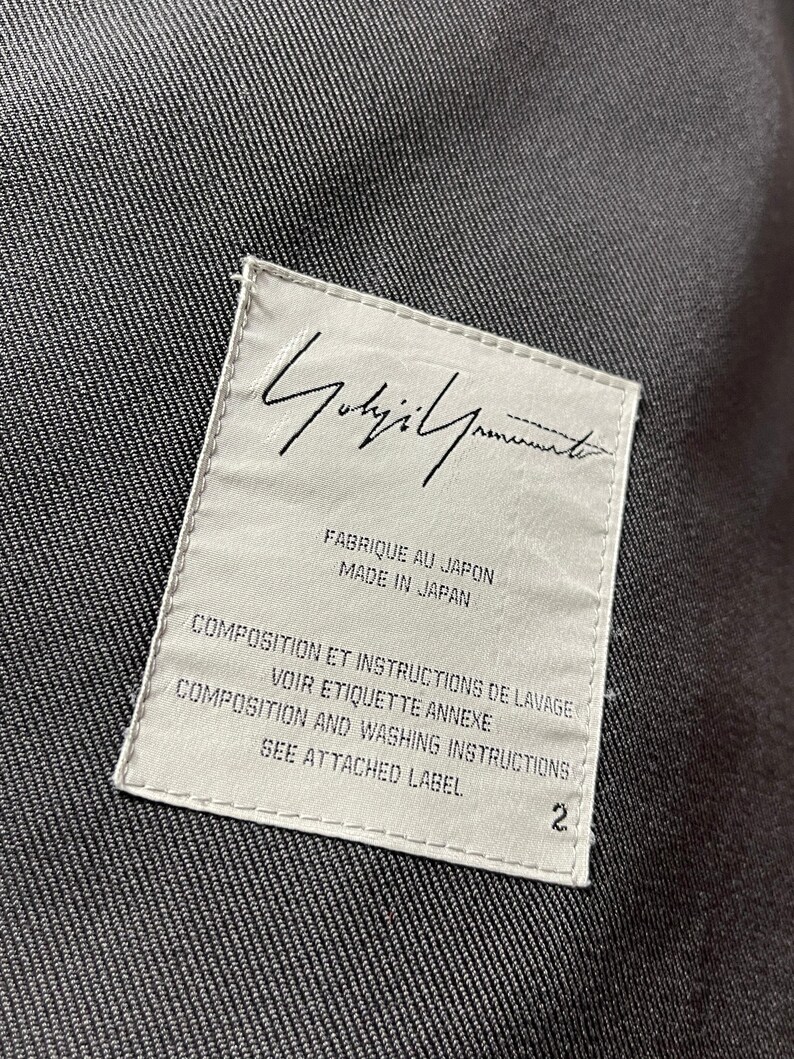 May include: A white label with black text, featuring the brand name "Yohji Yamamoto" in cursive script. The label includes text in English and French, indicating the item's origin and care instructions. The number "2" is printed on the label.