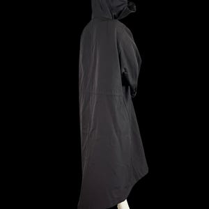 May include: A black hooded coat with a long, flowing design. The coat features a drawstring waist and a hood. The model is wearing the coat over a white outfit. The coat is made of a lightweight material and is suitable for various weather conditions.