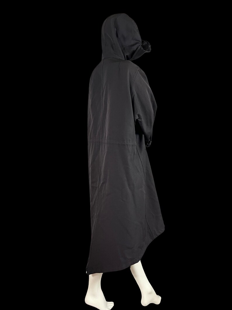 May include: A black hooded coat with a long, flowing design. The coat features a drawstring waist and a hood. The model is wearing the coat over a white outfit. The coat is made of a lightweight material and is suitable for various weather conditions.
