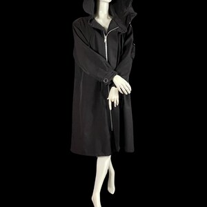 May include: A long black hooded coat with a silver zipper closure. The coat has long sleeves and a button detail on the cuffs. The mannequin is wearing white gloves and is set against a black background.